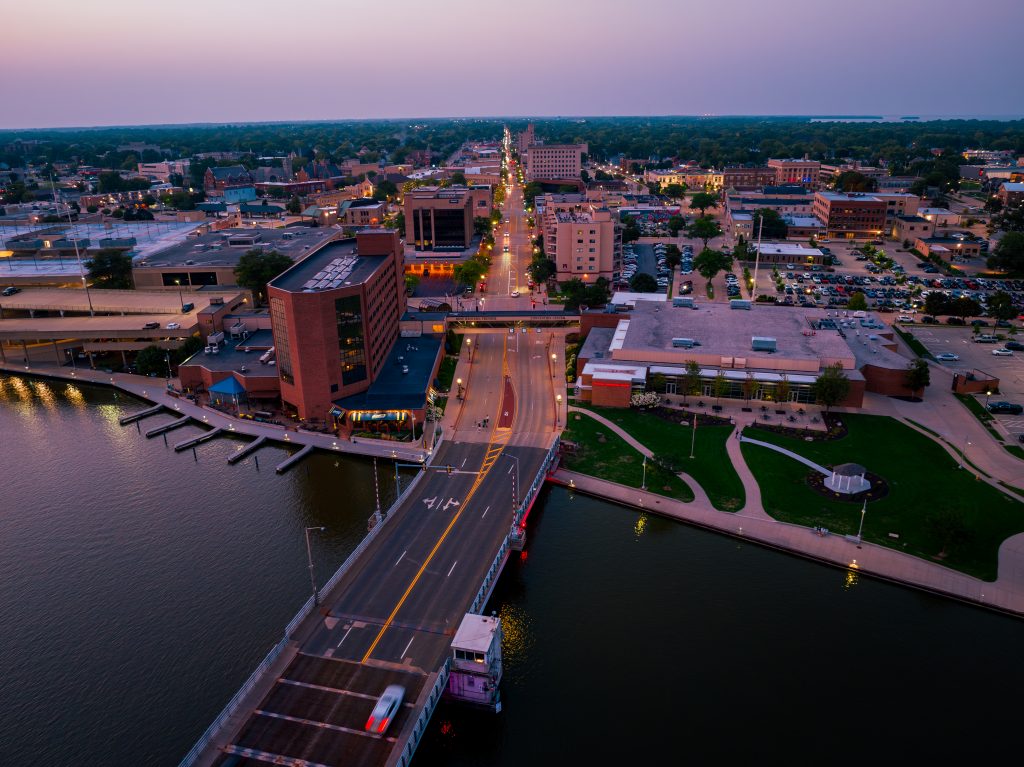 Aerial view of downtown Oshkosh, Wisconsin at dusk showing the riverfront, bridge, and illuminated city streets.